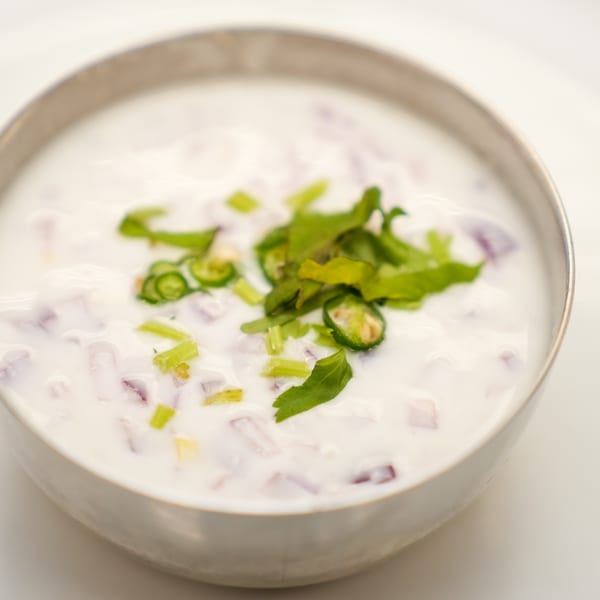 A silver bowl of raita, a yogurt-based condiment, with chopped red onions, green chilies, and cilantro.