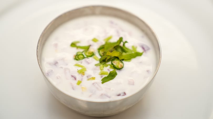 A silver bowl of raita, a yogurt-based condiment, with chopped red onions, green chilies, and cilantro.
