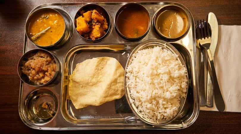A metal thali tray with various Indian dishes, including rice, curries, and papadum.