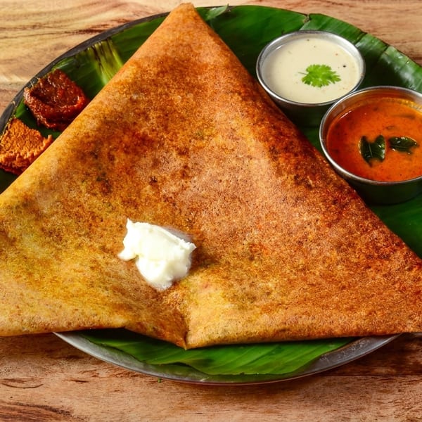Golden-brown dosa with a dollop of butter, served with two chutneys and a sambar on a banana leaf.