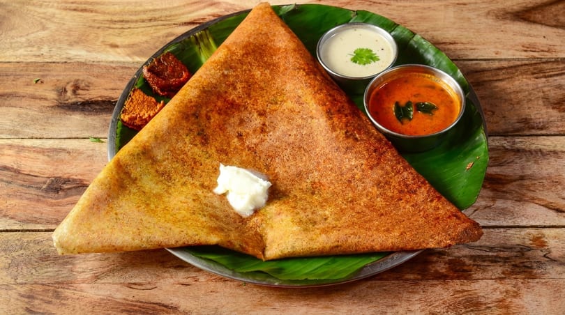 Golden-brown dosa with a dollop of butter, served with two chutneys and a sambar on a banana leaf.