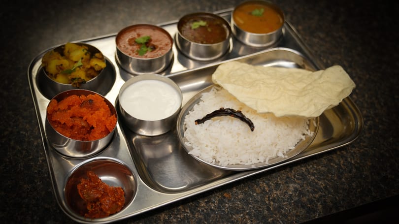 A stainless steel thali tray with various Indian dishes including rice, curries, and sweets.